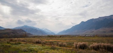 panoramic view of a valley with a mixture of dry and green vegetation, utility poles, and sparse buildings under a cloudy sky. Snow-capped mountains loom in the distance