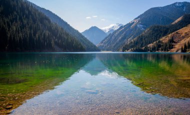 a serene alpine lake with crystal-clear waters reflecting the sky, bordered by dense coniferous forests and a backdrop of snow-dusted mountains under a blue sky
