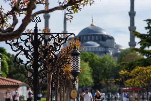 stock image Ornate wrought iron lamp posts and budding trees line a path with the iconic silhouette of a historical mosque's minaret and dome in the background