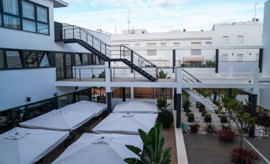 modern building courtyard, characterized by clean lines and a monochrome color palette. A staircase connects the different levels, while parasol-covered tables suggest a dining or relaxation area clipart