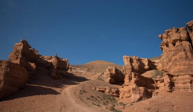 Sakin fotoğraf, sıcak ve güneşli bir günde yakalanan engin mavi gökyüzünün altında yükselen kırmızı kumtaşı oluşumlarıyla çöl kanyonunun huzurlu güzelliğini sergiliyor.