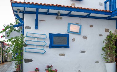 Pencere çerçeveleri, kepenkler ve yön okları gibi mavi boyanmış ahşap detayların yer aldığı beyaz bir duvar, Yunan ada mimarisinin tipik özellikleriyle çiçek açan bitkilerin bir zeminine kurulmuş..