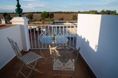 İşlenmiş demir mobilyalar ve dekoratif bir masa ile sakin bir balkon, berrak bir mavi gökyüzü altında sakin bir manzara, rahatlatıcı bir Akdeniz ayarı özünü somutlaştırır.