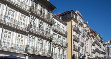 Süslü demirden balkonları, çizgili cepheleri ve açık mavi gökyüzü altında belirgin mimari detayları olan zarif, tarihi Avrupa binaları..