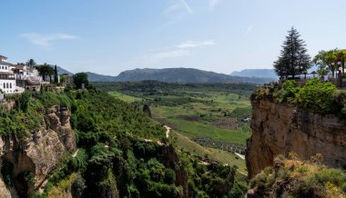 White-washed buildings crown a rugged cliff, overlooking a tapestry of green fields and distant mountains, under a bright azure sky. clipart