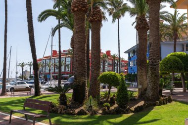 Uzun palmiye ağaçları, kırmızı ve mavi cepheli renkli binalar ve açık mavi gökyüzünün altında arka planda yelkenli tekneleri olan bir marina ile dolu sakin bir deniz gezisi.