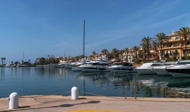 Palmiye ağaçları, demirlemiş yatları ve berrak mavi gökyüzünün altında teraslarla kaplı zarif binaları olan lüks bir rıhtım.