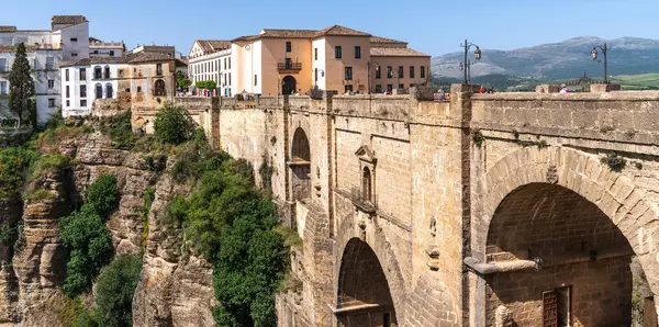 Stock image An ancient stone bridge arches majestically over a chasm, connecting two parts of an old town Ronda against a backdrop of rolling hills and clear skies.