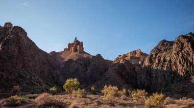 Berrak bir mavi gökyüzü altında engebeli, güneşli uçurumlarla çevrili kurak vadi tabanını beneklendiren yemyeşil çalılarla dolu sakin bir çöl manzarası gözler önüne seriliyor.