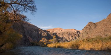 Amber foliage frames a mountains river, meandering at the foot of towering, rugged cliffs under the gentle embrace of a clear azure sky. clipart