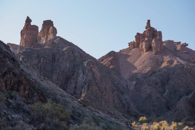 Görkemli kumtaşı oluşumları yumuşak bir gökyüzüne karşı keskin bir şekilde yükselirken, yürüyüşçüler bu engebeli çöl manzarasının ihtişamına ölçek ekliyorlar..