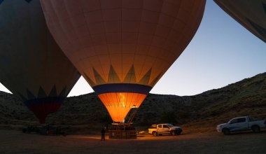 Şafak sökerken, şafak öncesi ışığa karşı sıcak hava balonları parlıyor, engebeli arazinin üzerindeki engin, açık gökyüzüne yükselmeye hazırlanıyorlar..