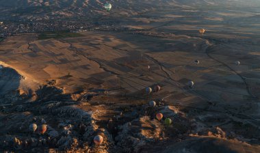 Şafakta, sıcak hava balonları dalgalanan dağların gölgelediği geniş bir vadinin üzerinde süzülür. Renkleri solan geceye karşı canlıdır. Aşağıda, yama alanları ve uzak kasabalar.