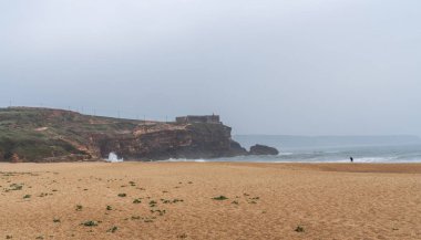 Altından kumlu sisli bir sahil manzarası, tepesinde bir yapı olan engebeli bir uçurum, ve kıyı şeridi boyunca dağılmış figürler sessiz, bulutlu bir gökyüzünün altında.