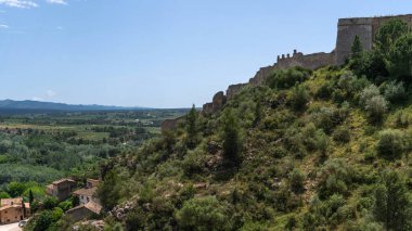 Antik kalenin geniş manzaralı harabeleri yeşil bir yamacın tepesinde, açık bir gökyüzü altında yamalı arazileri olan geniş bir vadiye bakıyor.
