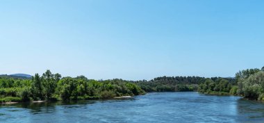 A tranquil river cuts through lush foliage under an expansive blue sky, with distant hills rolling into the horizon, embodying serene natural beauty. clipart