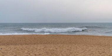 Kasvetli bir gökyüzünün altında dalgalarla buluşan altın kumlarla dolu sakin bir sahile bakıyor. Güçlü dalgalar kumlu sahile doğru yükseliyor. Huzur ve yalnızlık hissi veriyor..