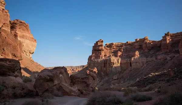 stock image A desert canyon landscape basks in soft sunlight, with towering red sandstone cliffs flanking a winding dirt path, evoking a sense of solitude and natural grandeur.