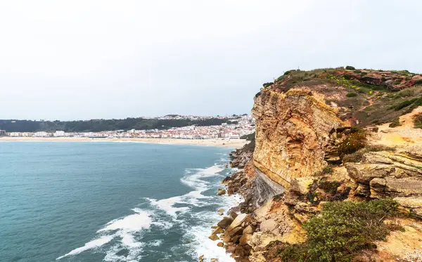 stock image a stunning coastal view from atop a rugged cliff, overlooking a long stretch of sandy beach that reaches towards a quaint seaside town, under a muted sky.