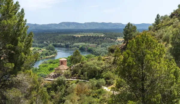 stock image Panoramic view of a serene river meandering through a lush valley, with a solitary chapel perched on the hillside and mountains in the backdrop