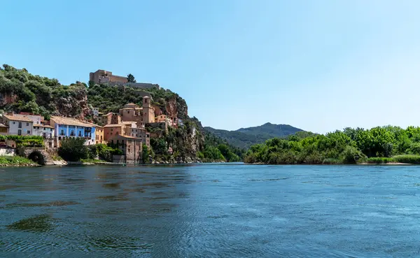 stock image a picturesque riverside village Miravet, Spain. Ancient buildings clinging to the steep hillside, the river flows calmly in the foreground, framed by the lush greenery of the surrounding landscape.