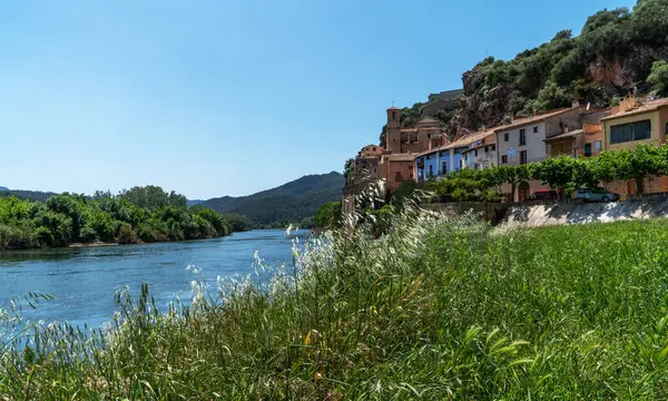 stock image a serene river flowing past a hilltop village, with the architecture of the village merging seamlessly into the lush, natural landscape under a clear blue sky