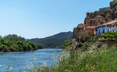 a serene river flowing past a hilltop village, with the architecture of the village merging seamlessly into the lush, natural landscape under a clear blue sky clipart