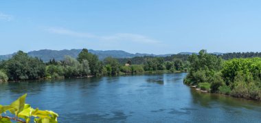 A calm river meanders through a verdant landscape, flanked by lush trees and set against a backdrop of gentle mountain ridges under a clear blue sky. clipart