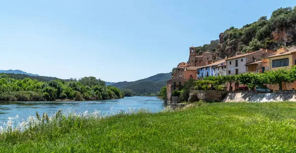 stock image A serene river gently flows by a historic village with terracotta roofs, nestled against a backdrop of lush hills under a clear blue sky, capturing the essence of rural tranquility