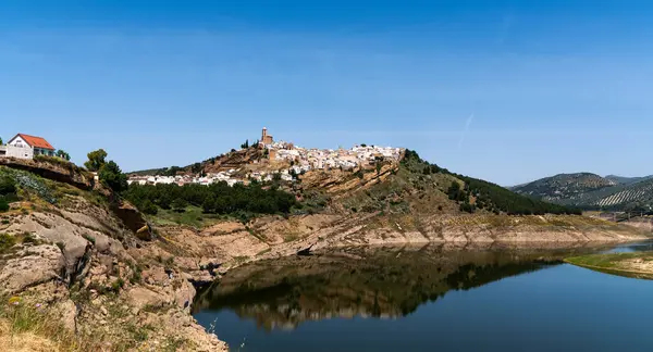 stock image picturesque landscape a hilltop Spanish village with clustered white houses and a church, reflected in the tranquil waters of a foreground reservoir, a backdrop of clear blue skies and hills.