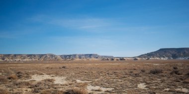 Kazakistan 'daki Ustyurt Platosu' nun kurak manzarası. Çorak ovalar, berrak mavi bir gökyüzünün altındaki aşınmış uçurumlara doğru uzanır. Nadir bitki örtüsü toprağı kaplar.