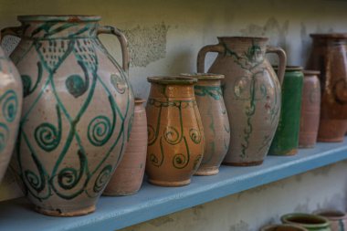 Mavi ahşap raflarda yıpranmış bir duvara karşı seramik sürahi ve çömlek koleksiyonu. Çömlekçilik geleneksel zanaatkarlığı yansıtıyor ve ayara kırsal bir cazibe katıyor..