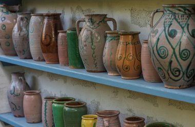 Mavi ahşap raflarda yıpranmış bir duvara karşı seramik sürahi ve çömlek koleksiyonu. Çömlekçilik geleneksel zanaatkarlığı yansıtıyor ve ayara kırsal bir cazibe katıyor..