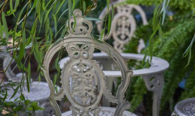 An old ornate, white wrought-iron garden chair surrounded by greenery. The intricate design of the chair back is partially obscured by hanging leaves, creating a tranquil and vintage atmosphere. clipart