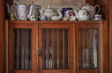 An antique wooden cabinet with an array of decorative porcelain teapots. The cabinet has glass-paneled doors, the intricate details of the porcelain collection, creating a warm, vintage atmosphere. clipart