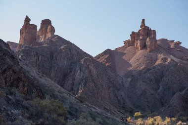 Görkemli kumtaşı oluşumları yumuşak bir gökyüzüne karşı keskin bir şekilde yükselirken, yürüyüşçüler bu engebeli çöl manzarasının ihtişamına ölçek ekliyorlar..
