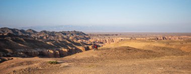 Uçsuz bucaksız gökyüzünün altında geniş bir çöl manzarası var. Gezginler, kayan tepeler ve katmanlı kanyonların kurak arazisinde karla kaplı uzak dağlar sayesinde görkemli bir zemin oluşturuyor..