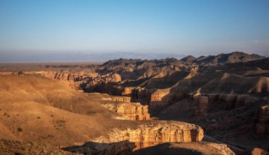 Batan güneşin sıcacık parıltısı engin bir kanyonun katmanlı kaya oluşumlarını vurguluyor. Gölgeler dalgalanan araziye yayılıyor ve uzak karlı dağlara doğru yol alıyor..
