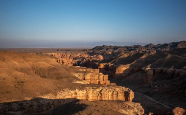 Batan güneşin sıcacık parıltısı engin bir kanyonun katmanlı kaya oluşumlarını vurguluyor. Gölgeler dalgalanan araziye yayılıyor ve uzak karlı dağlara doğru yol alıyor..