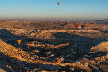 multiple hot air balloons rise above a stark landscape of eroded rock formations, casting colorful silhouettes against the clear sky. clipart