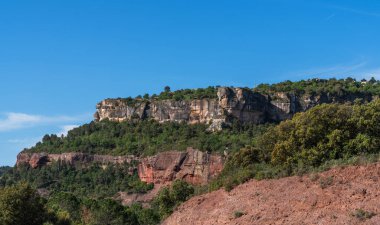Bereketli, kırmızımsı kahverengi önplanı ile berrak mavi gökyüzünün altında yükselen kayalıklarla taçlandırılmış, dingin bir manzara ortaya çıkar..