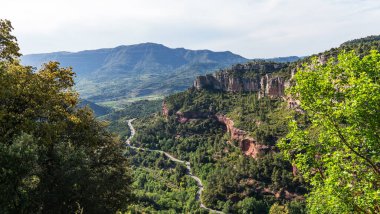 A winding road meanders through a verdant valley, flanked by dramatic red cliffs and lush forests, the of distant mountains against a bright sky. clipart