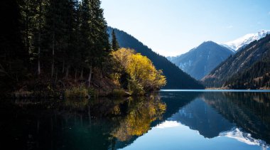 The alpine splendor of a mountain lake where the stillness of the water mirrors the surrounding forest and the grandeur of the snow-dusted peaks, under the vast expanse of a clear blue sky. clipart