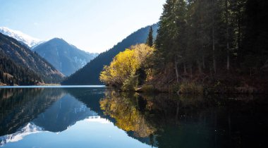 Bir dağ gölünün alp ihtişamı. Suyun durgunluğu etrafındaki ormanı yansıtıyor. Kar tozuyla kaplı tepelerin ihtişamı berrak mavi gökyüzünün uçsuz bucaksız genişliğinin altında..