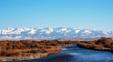 Huzurlu bir nehir engebeli bir arazide, sonbahar çalılıklarıyla çevrili, derin mavi gökyüzünün altındaki görkemli karla kaplı dağlara doğru ilerliyor.