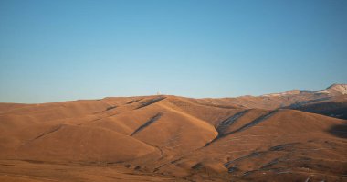 Altın saat ışığı dalgalanan tepeler ve kuru otlaklar üzerine uzun gölgeler düşürüyor sakin, yüksek irtifada dağ manzarası.