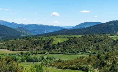 Ağaçlarla kaplı yeşil bir vadiyi kaplayan panoramik bir manzara, engin mavi gökyüzünün altında kayan tepelere ve uzak dağlara doğru süpürüyor..