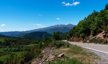Dolambaçlı bir yol yamacın yamacını sarar, yemyeşil vadiler ve tepeler boyunca manzaralı bir yolculuk sunar, engin, bulutlu gökyüzünün altındaki görkemli dağ sırasına götürür..