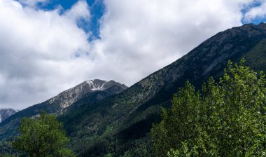 hala karla kaplanmış, çarpıcı, görkemli dağ zirveleri, yemyeşil ormanların üzerinde, bulutlarla benekli dinamik bir gökyüzü altında, dağlık bir manzaranın ihtişamını yakalıyor.