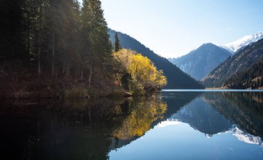 Bir dağ gölünün alp ihtişamı. Suyun durgunluğu etrafındaki ormanı yansıtıyor. Kar tozuyla kaplı tepelerin ihtişamı berrak mavi gökyüzünün uçsuz bucaksız genişliğinin altında..